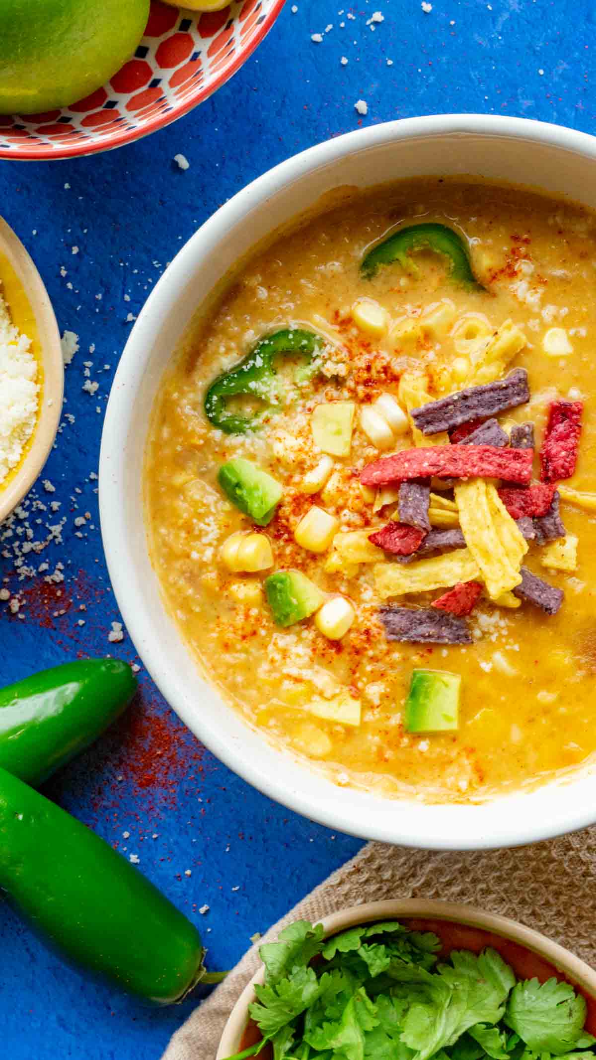 Mexican Street Corn Soup in a white bowl with a blue countertop and fresh vegetables surrounding it